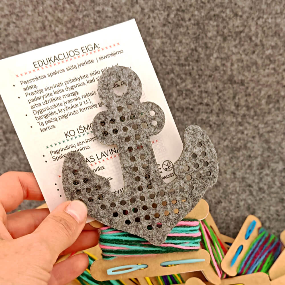 Nesiuvinėta veltinio inkaro formelė virš gulinčios „Edukacijos eiga“ instrukcijos kortelės ir žalių gijų.
