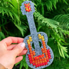 Žalių augalų fone rankoje laikoma pilnai išsiuvinėta pilko veltinio gitara su ryškiais geltonais, oranžiniais ir raudonais kryžminiais dygsniais korpuse.